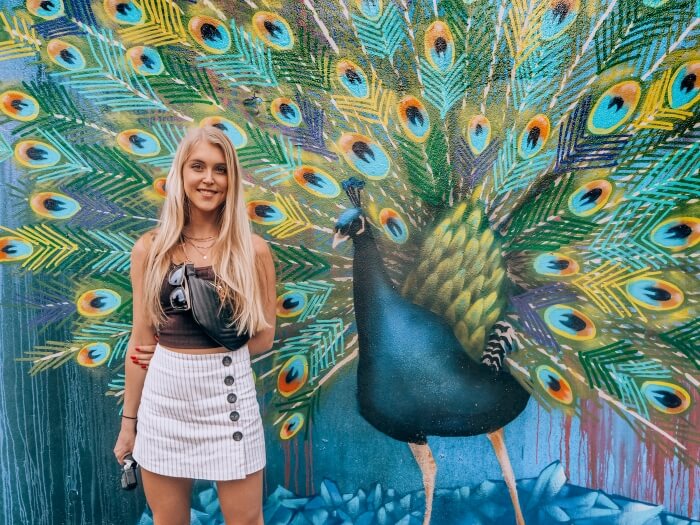a girl standing in front of a colorful mural depicting a peacock in Beco do Batman, a street art gallery in Sao Paulo