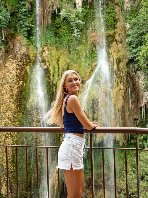 Me leaning on a railing with the Cascade de Sillans waterfall and its lush vegetation in the background