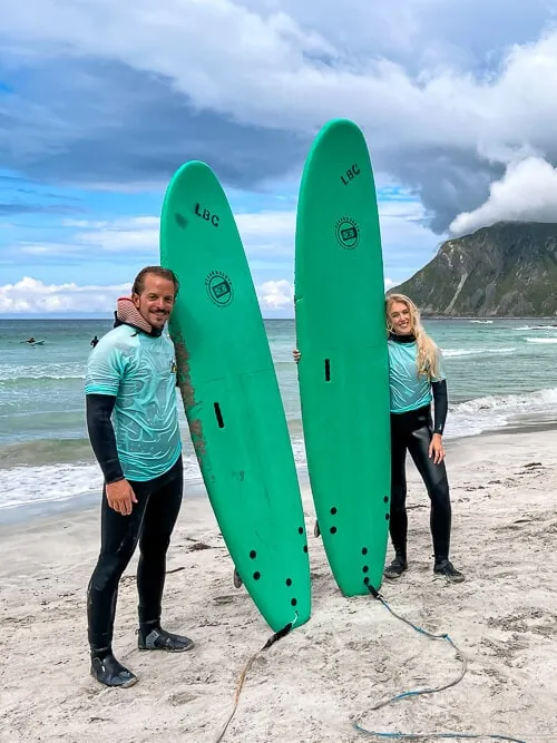 Me and my boyfriend in wetsuits standing on a sandy beach holding our green surf boards.