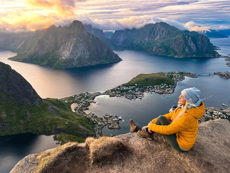 Me in a yellow jacket sitting on a rock on top of Reinebringen overlooking a scenic fjord surrounded by mountains. 