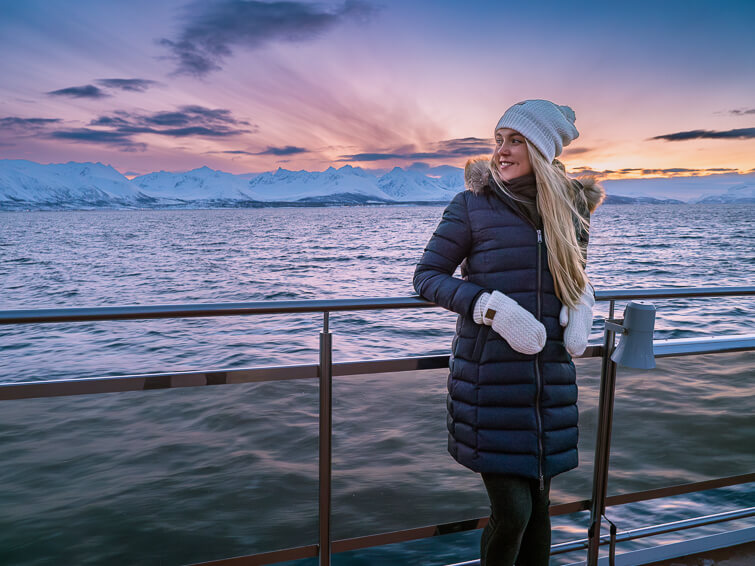 Me posing on the outdoor deck of a boat with picturesque Arctic landscapes and colorful sky in the background.