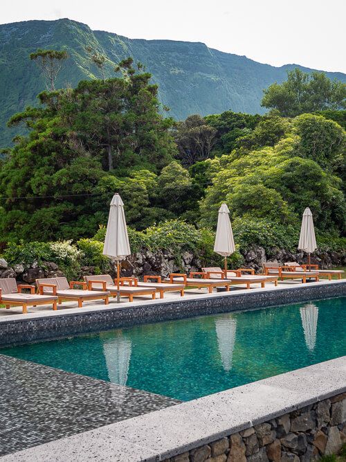 A turquoise swimming pool surrounded by sunbeds and tall green mountains in the background