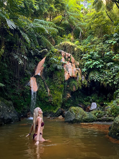 Me standing in a natural pool at Caldeira Velha hot springs with a small waterfall and lush green plants in the background.