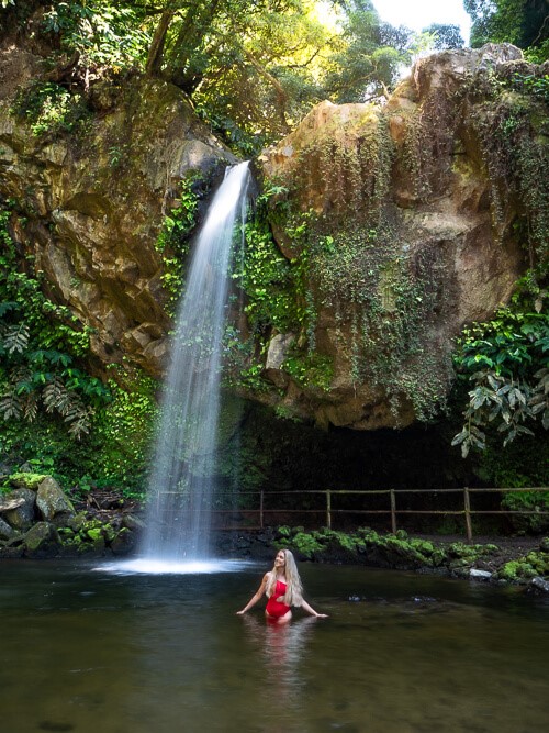 Me posing in front of Cascata da Gruta waterfall cascading down a rocky cliff covered with hanging plants.