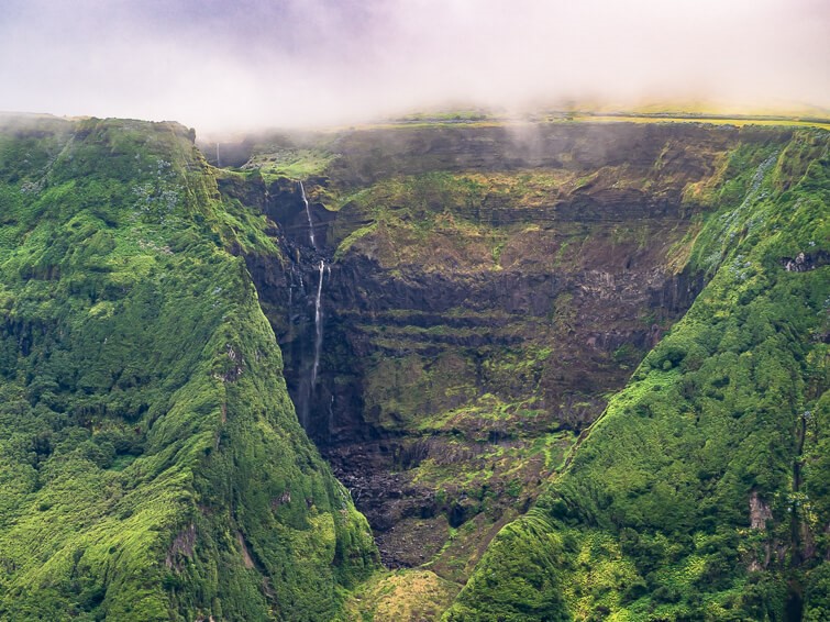 Thin stream of water of Cascata da Ribeira Grande dropping down a vertical cliff surrounded by green mountains.