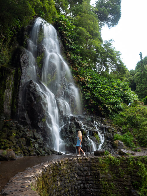 Me walking in front of Cascata da Ribeira dos Caldeirões waterfall flowing down a rugged mountainside, surrounded by lush foliage.