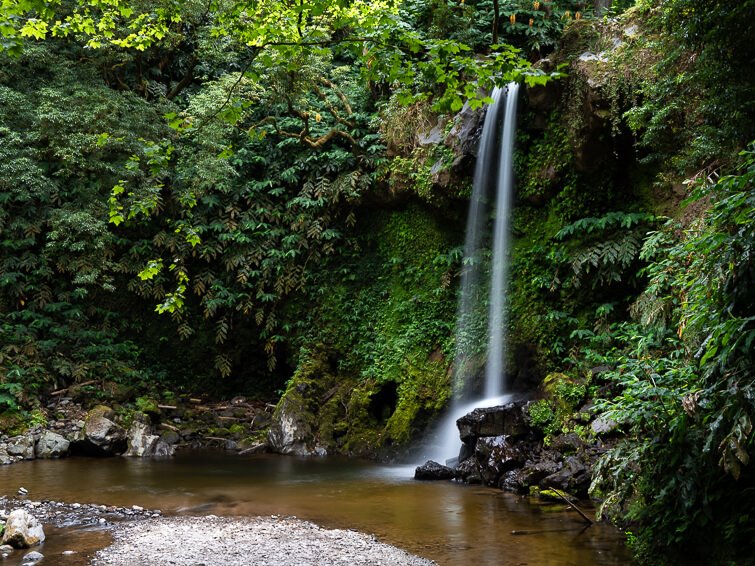 A thin stream of water of Cascata do Teofilo waterfall dropping down from a rock into a natural pool surrounded by dense green vegetation.