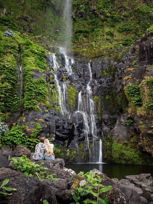 Me and my husband sitting on a rock and looking at Cascata do Poço do Bacalhau waterfall gently cascading down black rocks.