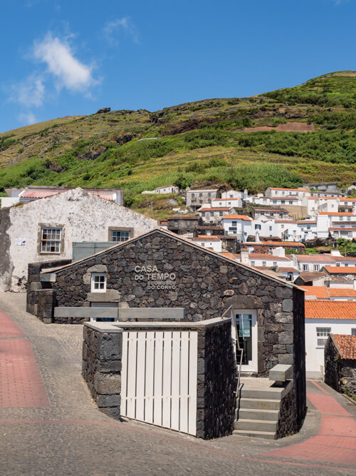 The small Casa do Tempo museum made of black volcanic rocks and the white houses of Vila do Corvo in the background.