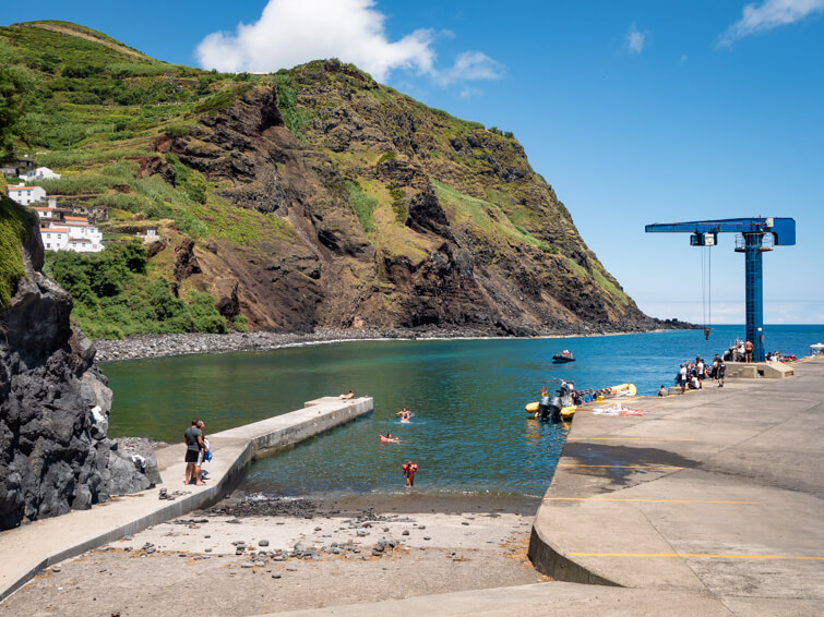 People swimming in the turquoise water at the port of Corvo, with a rugged green mountain in the background.