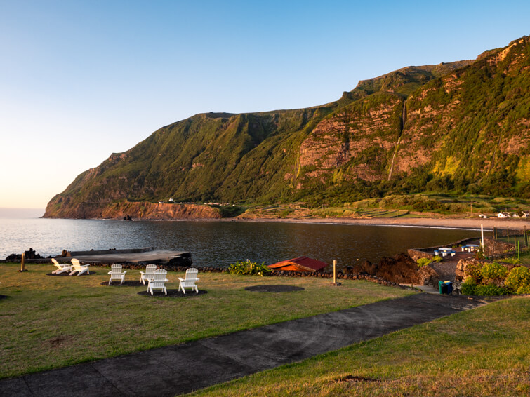 The scenic waterfront of Fajã Grande village during sunset with tall green mountains as a backdrop.