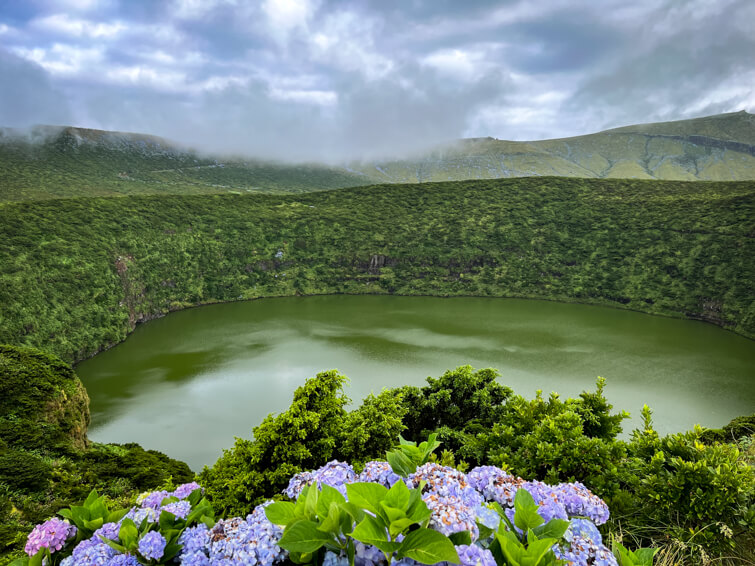 The small round Lagoa Negra crater lake surrounded by steep green slopes and purple hydrangea flowers.