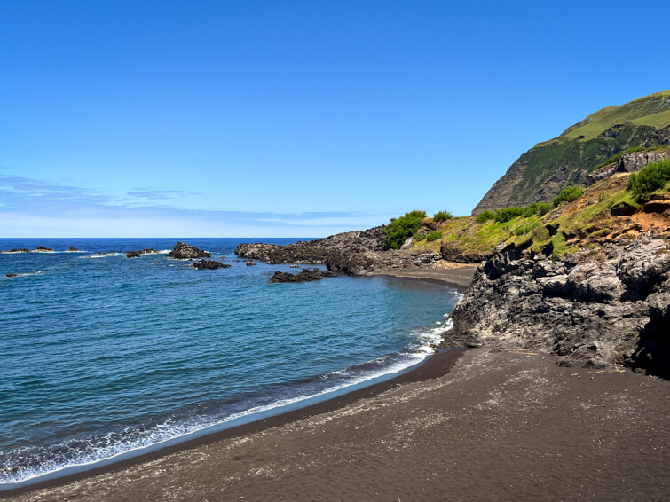 Praia da Areia Beach with its dark volcanic sand, blue water, and green mountains in the background.