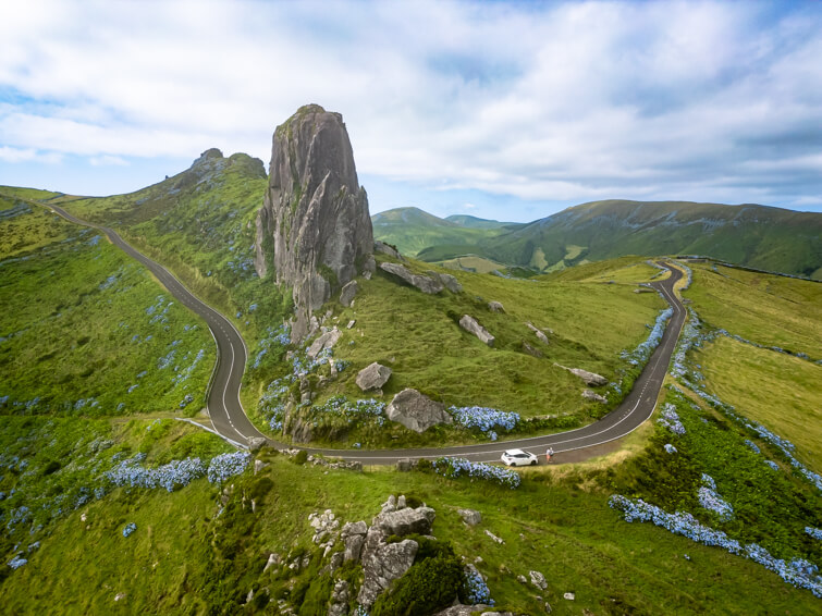 A small road meandering through green pastures and around the towering rock formation of Rocha dos Frades.